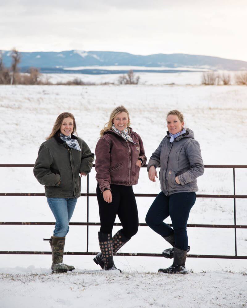 The three women of Montana RancHERS Beef Co standing against a metal fence in the winter.